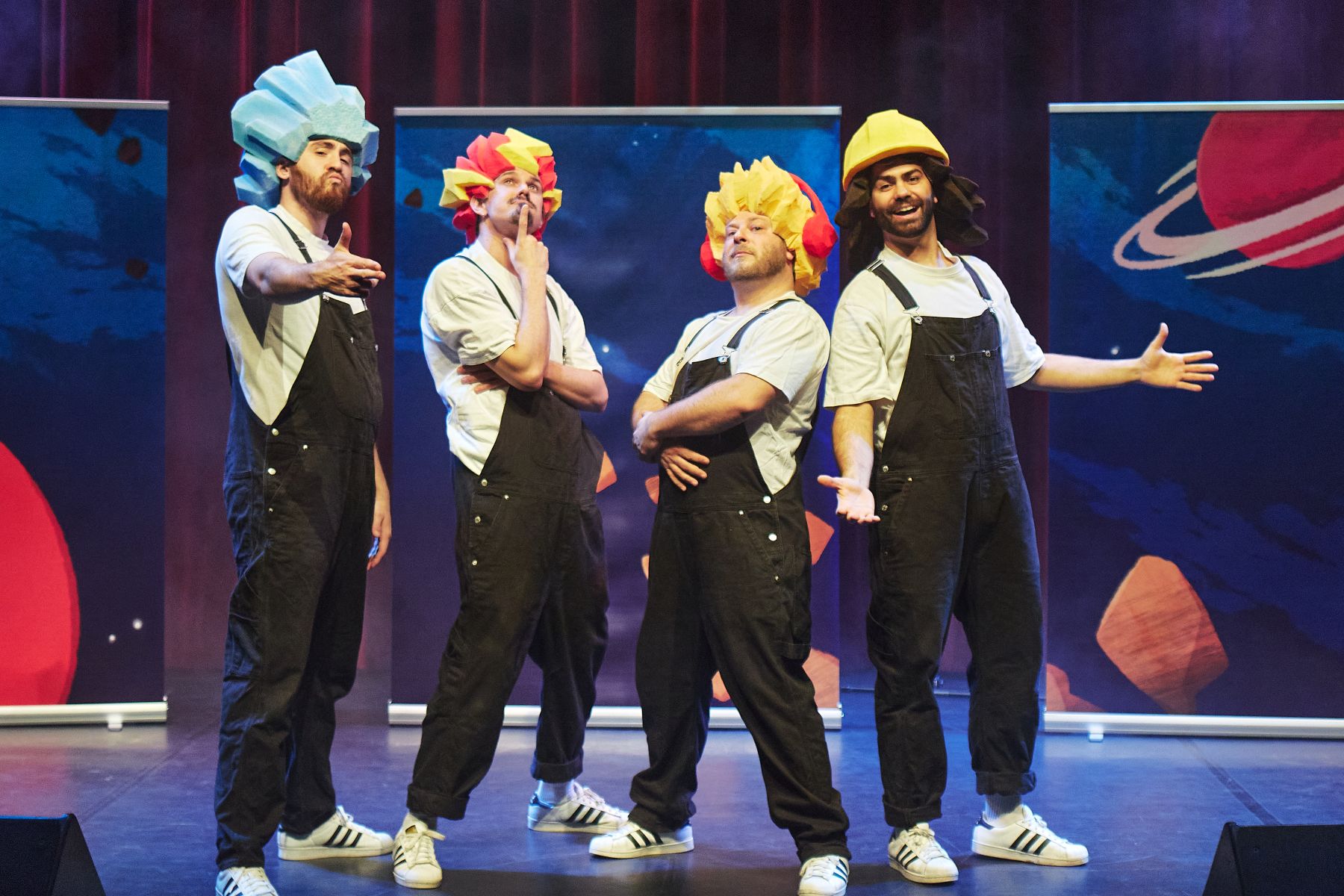 Das Bild zeigt die Razzelbande, in lustigen Posen, auf einer farbenfrohen Bühne mit Weltraumthema. Sie tragen alle schwarze Latzhosen und weiße Pullover. Sie tragen bunte große Perücken aus Kunststoff.