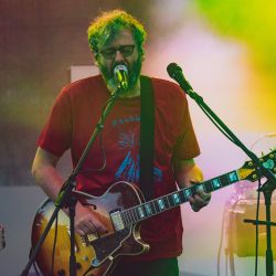 Das Bild zeigt einen Mann mit Brille, welcher Gitarre spielt und dazu singt. Er trägt ein rotes Oberteil mit blauer Aufschrift.