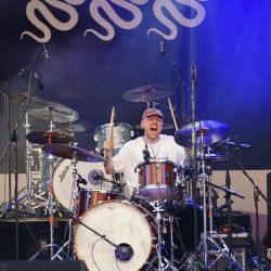 Schlagzeuger in Action. Er hält beide Drumsticks nach oben und sein Mund ist weit aufgerissen, so als würde er mitsingen.