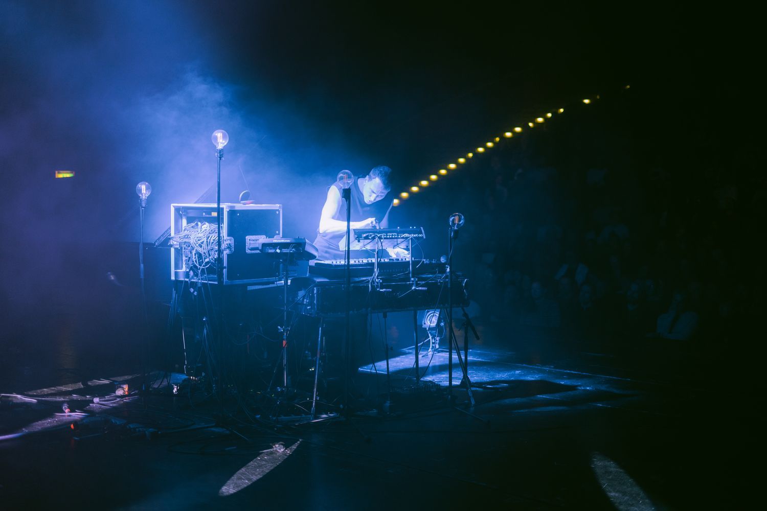 Auf dem Foto ist Martin Kohlstedt während eines Auftritts zu sehen. Umgeben von blauem Licht und leichtem Nebel steht er an seinem DJ-Pult und E-Piano.