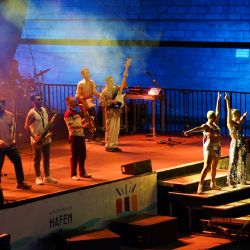 Dieses Bild ist seitlich von der Bühne aufgenommen. Die Band scheint sich zu verabschieden, Die zwei Sängerinnen posieren auf der Treppe, die restlichen 6 Musiker stehen mit ihren Instrumenten auf der Bühne.