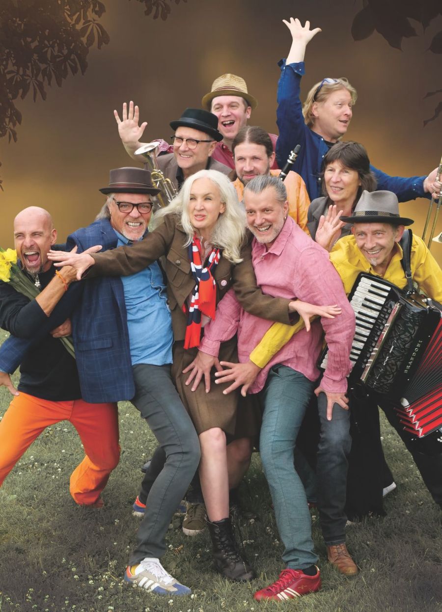 Das Bild zeigt eine lebhafte und fröhliche Gruppe von Personen, die ausgelassen posieren. Alle Personen strahlen Energie und Freude aus, wobei sie in bunter Kleidung gekleidet sind. Im Vordergrund hält jemand gelbe Blumen, andere Instrumente.