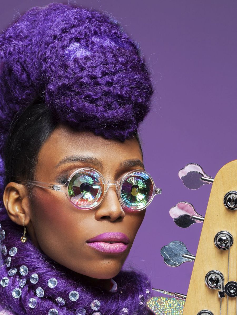 Das Porträt zeigt Nik West mit einer auffälligen Frisur aus voluminösen, lila gefärbten Haaren, passend zu einem violetten Hintergrund. Sie trägt eine runde, reflektierende Brille, die das Licht in bunten Farben bricht. Ihr Make-up ist kräftig.