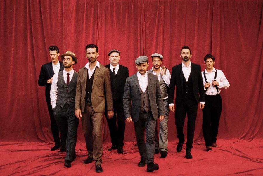 Auf dem Bild sind die Musiker des Babylon Circus zu sehen, wie sie vor einem roten Vorhang stehen und es wirkt als würden sie auf die Kamera zulaufen. Sie tragen Anzüge und teilweise Hüte im Stil der 30er Jahre.