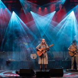 Das Bild zeigt eine Band auf der Bühne, der Hintergrund ist rot beleuchtet aber mit weiteren Scheinwerferm werden blaue Lichtkegel auf die Band gestrahlt.