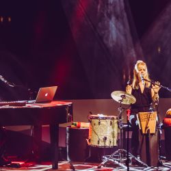 Das Bild zeigt links einen Mann welcher an einem Flügel sitzt. Rechts vom Flügel stehen viele Intrumente, vor allem Trommeln. Mitten drin sitzt eine Frau welche grade in ein Mikrofon singt.