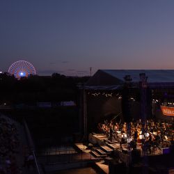 Das Bild zeigt eine Aufnahme der Bühne von oben. Es ist bereits dunkel. Das Orchester ist jecoh hell beleuchtet. Im Hintergrund sieht man beleuchtete Fahrgeschäfte des Volksfestes.