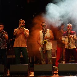 Das Bild zeigt die Band in einem sehr dunklen Licht mit Nebel. Alle 4 blicken nach vorne und spielen ein Blasinstrument. Im Hintergrund ist ein weiterer Mann versteckt.