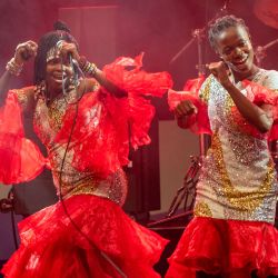 Das Bild zeigt zwei Personen, welche tanzen. Beide tragen auffällige rot-goldene Kleidung mit sehr viel Glitzer.