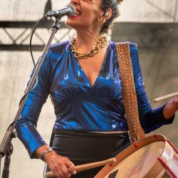 Ayom hat eine Trommel umhängen, auf welcher sie spielt und sind dabei in ein Mikrofon.