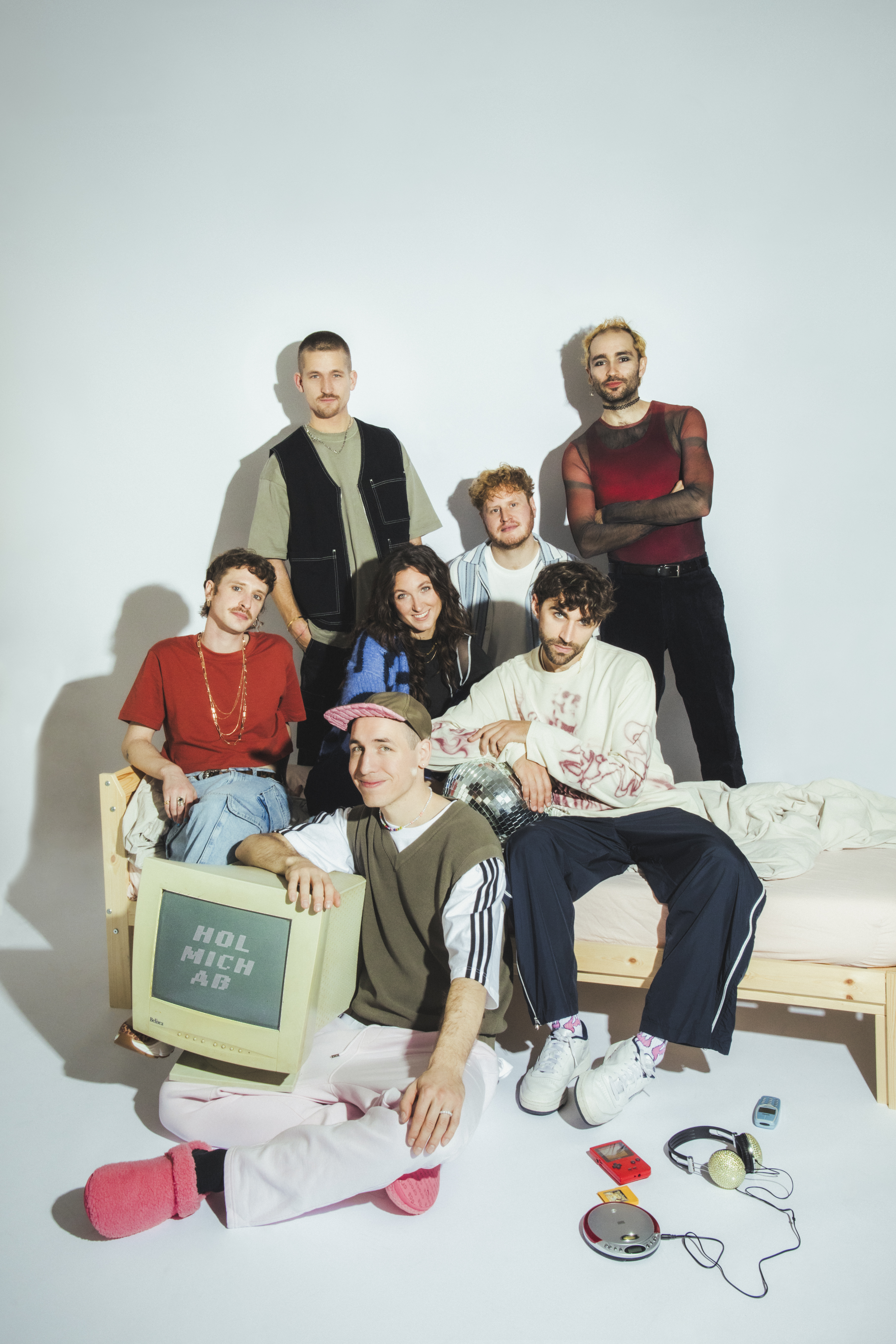 Die Bandmitglieder der Band Make a Move posieren zusammen auf, bzw. bei einem Bett. Vier sitzen auf dem Bett, einer davor. Zwei stehen dahinter. Sie tragen bunte Kleidung und lächeln. Auf dem Schoß des Sitzenden liegt ein Röhrenfernseher.
